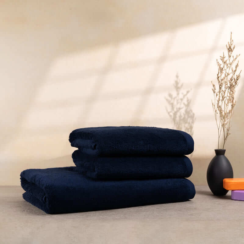 Set aus 3 dunkelblauen Handtüchern, auf einer neutralen Oberfläche mit Vase und Deko-Elementen gestapelt.