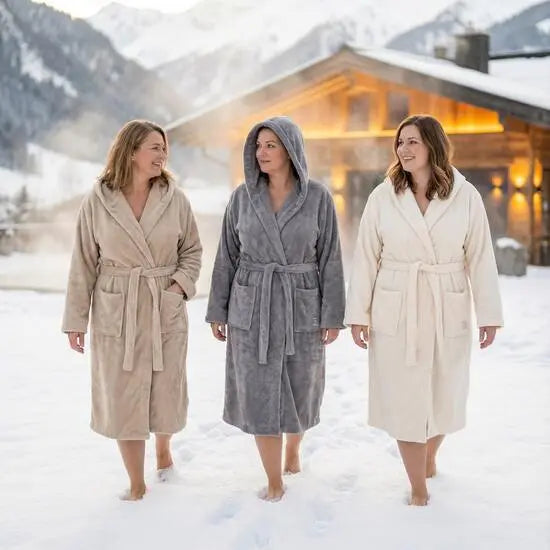 Drei Frauen in Bomlins Bademänteln in XXL aus GIZA Baumwolle in den Farben Cashmere Beige Grau Naturweiß gehen barfuß durch den Schnee vor einer Berghütte.
