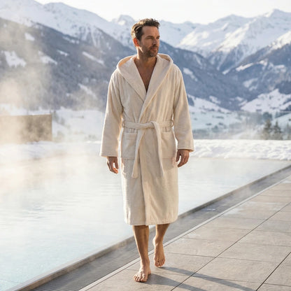 Mann in beigen Bomlins Bademantel aus GIZA Baumwolle geht barfuß am beheizten Außenpool vor winterlicher Bergkulisse.